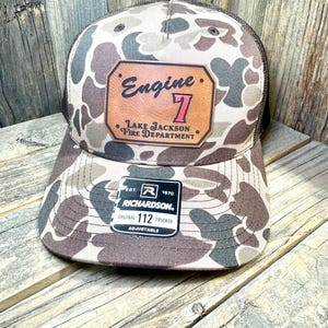 May include: A camouflage trucker hat featuring a brown leather patch with the text Engine 7 and Lake Jackson Fire Department. The hat has a brown mesh back and a black Richardson 112 sticker on the brim. The pattern consists of brown, tan, and dark green shapes.
