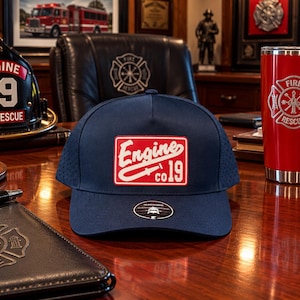 May include: A navy blue baseball cap featuring a rectangular red patch on the front with white text that reads Engine co 19 and a graphic of a fire hose nozzle. The cap has a perforated side panel and a small circular sticker on the brim.