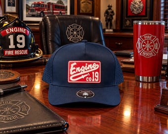 May include: A navy blue baseball cap featuring a rectangular red patch on the front with white text that reads Engine co 19 and a graphic of a fire hose nozzle. The cap has a perforated side panel and a small circular sticker on the brim.