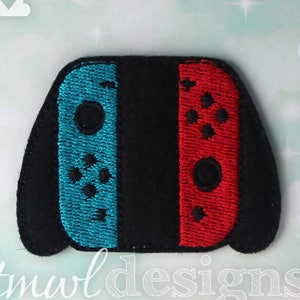 May include: Embroidered patch in the shape of a video game controller. The controller has a black center with a blue left side and a red right side. The text "tmwl designs" is at the bottom.