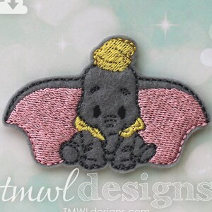 May include: A gray felt embroidered patch of Dumbo the elephant from Disney. Dumbo is sitting down with his ears out and wearing a yellow hat. The patch is pink and gray.