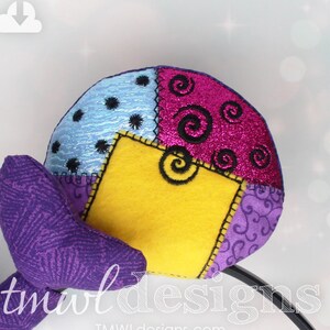 Puede incluir: Diadema de patchwork de tela morada, azul, amarilla y rosa con un diseño de espiral. La diadema está hecha de fieltro y tiene un diseño de espiral bordado en negro. La diadema está unida a una diadema negra.