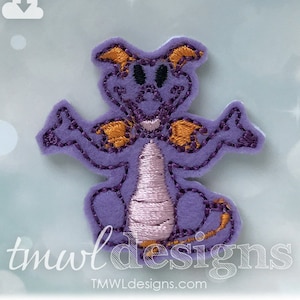 May include: Purple felt applique of a cartoon dragon with orange wings and black eyes. The dragon is sitting with its arms outstretched.