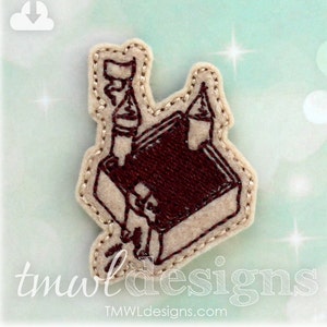 Puede incluir: Una aplicación de fieltro marrón de un castillo con un contorno blanco. El castillo tiene dos torres y una bandera en la parte superior. La aplicación está sobre un fondo azul claro con purpurina blanca.