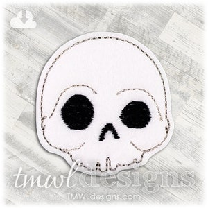 May include: White skull-shaped patch with black eye sockets and nose. The patch has a silver stitched outline and is on a white background. The text "tmwl designs" and "tmwldesigns.com" are at the bottom. A Halloween-themed item.