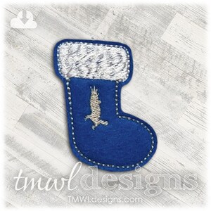 Puede incluir: Una media de fieltro azul con un puño de fieltro blanco y un pingüino de fieltro blanco en su interior. La media está decorada con costuras blancas.