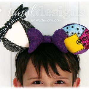 May include: A purple bow headband with two fabric ears. One ear is black and white striped with a black bat wing design. The other ear is a patchwork of blue, yellow, pink, and white fabric with a swirl design.