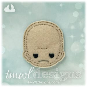 May include: A beige felt applique in the shape of a baby's face with black eyes and a stitched mouth. The applique has a stitched border. The text "tmwl designs" is at the bottom of the image.
