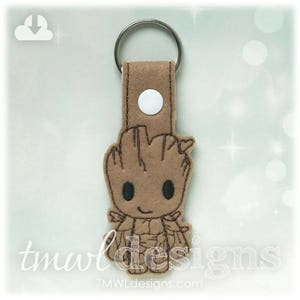 May include: A brown felt keychain featuring a cartoon character with a brown body, black eyes, and a brown smile. The character is wearing a brown felt outfit with a leafy design. The keychain has a white metal ring.