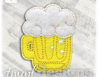 May include: A yellow felt beer mug with white foam, stitched with tan thread. The mug has vertical stitched lines and small white starburst details. The text "tmwl designs" and "tmwldesigns.com" are at the bottom.