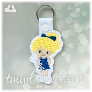 May include: A white keychain with a blue and yellow embroidered image of a girl holding a baton. The girl has blonde hair and is wearing a blue and white uniform. The keychain has a silver ring.