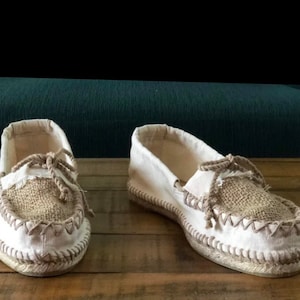 May include: Pair of off-white espadrille-style shoes with jute accents. The shoes feature a canvas upper, jute toe, and rope detailing. The soles are made of woven material, and the shoes are designed with a moccasin-style construction.