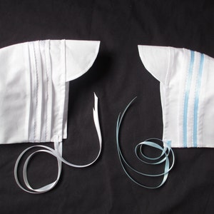 May include: Two white bonnets with ties. One bonnet has a plain white tie, the other has a light blue tie.
