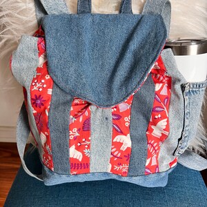 May include: A denim backpack with a red floral and bird pattern on the front flap and center panel. The backpack has a top handle, adjustable straps, and a side pocket for a water bottle. The bag is made of blue denim.