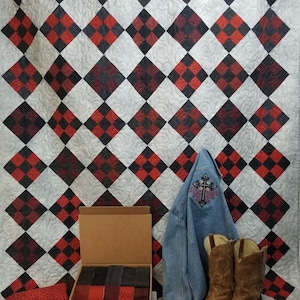 Könnte beinhalten: Eine handgefertigte Steppdecke mit einem sich wiederholenden Muster aus roten, schwarzen und weißen Rauten. Eine Jeansjacke mit Kreuzdesign, braune Cowboy-Stiefel und eine Schachtel mit Stoffquadraten werden vor der Steppdecke präsentiert.