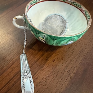 May include: A tea infuser with a silver-toned metal mesh ball and a decorative floral embossed handle attached by a chain, resting inside a white ceramic teacup featuring a green exterior band with painted pink and green floral motifs.