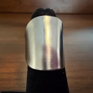 May include: A wide, curved silver-toned ring with a smooth, brushed metallic finish. The band features a solid, continuous surface that wraps around, showcasing a reflective sheen and a clean, unadorned design with no visible engravings or stones.