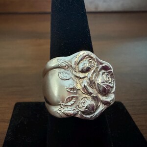 May include: A silver-toned ring crafted from a repurposed spoon, featuring a prominent embossed floral design of three roses with detailed petals and leaves. The band is wide and smooth, showcasing the original metallic luster and curved shape.