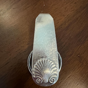 May include: A silver-toned metal money clip featuring a tapered rectangular shape with a rounded bottom. The lower portion of the clip is embossed with a detailed fan-shaped seashell motif, complete with radiating ridges and two small spiral accents.