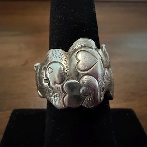 May include: A silver-toned metal ring featuring a raised, textured design of a cartoon bear character with heart-shaped details on its chest and face. The surface of the ring has a polished finish with engraved lines creating fur-like textures.