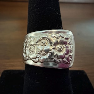 May include: A wide silver-toned ring featuring a raised, embossed floral pattern across the entire outer surface. The metal has a polished, reflective finish with detailed petals and leaves sculpted into the band, creating a textured appearance.
