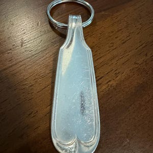 May include: A silver-colored metal keychain featuring a flat, elongated handle section from a piece of silverware. The handle has a smooth, reflective surface with subtle decorative grooved lines along the edges and a rounded, heart-shaped base.