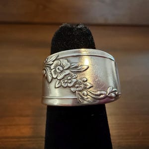 May include: A wide silver-toned metal ring featuring an embossed floral pattern that runs diagonally across the surface. The band has a smooth, polished finish with detailed engravings of leaves and small flowers, creating a textured appearance.
