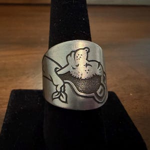Puede incluir: Un anillo ancho de metal en tono plateado con una ilustración grabada de un personaje de dibujos animados que se asemeja a un oso o criatura similar apoyando su cabeza sobre una superficie, con un relleno oscuro texturizado.