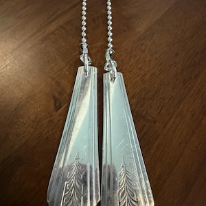 May include: Two silver-coloured fan pull chains with decorative, elongated pulls. Each pull features an etched design of a stylized tree or plant. The pulls are connected to the chains with small metal loops.