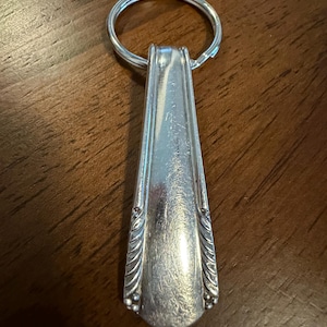 May include: A silver-toned metal keychain featuring a handle from a vintage spoon. The handle has a smooth, tapered center with decorative ribbed and beaded detailing along the lower edges, attached to a circular metal split ring.