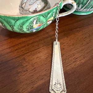 May include: A silver-toned metal tea infuser with a spherical mesh body and a decorative flat handle featuring an embossed floral pattern. The infuser is connected by a silver chain and rests inside a green and white patterned ceramic teacup.