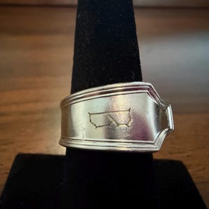 Puede incluir: Un anillo de plata hecho de una cuchara, con un diseño rectangular con un contorno estatal. El anillo se muestra sobre un soporte negro sobre un fondo marrón. La banda del anillo mide aproximadamente 1,9 cm de ancho.