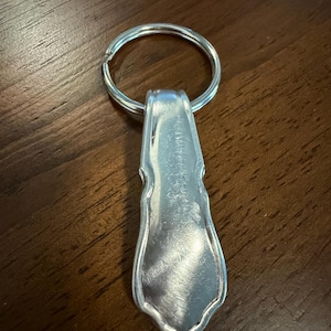Puede incluir: Un llavero de metal plateado que presenta un colgante plano, alargado y en forma de escudo con una superficie lisa y reflectante y un borde festoneado, unido a una anilla circular para las llaves.