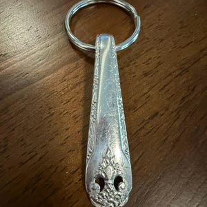 Puede incluir: Un llavero de metal plateado hecho con el mango de una cuchara vintage. El mango presenta un patrón floral y de hojas en relieve a lo largo de los bordes y un motivo decorativo detallado en la base, unido a una anilla metálica circular.