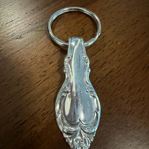 Puede incluir: Un llavero de metal plateado que presenta un mango decorativo recuperado de un cubierto. El mango tiene un patrón ornamental en relieve con volutas y está unido a una anilla circular para sujetar las llaves.