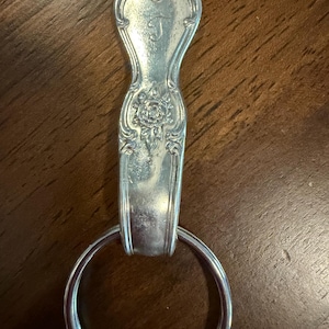 Puede incluir: Un llavero de metal plateado que presenta un mango de cuchara reutilizado como componente principal. El mango muestra un patrón floral en relieve y la letra J grabada en la parte superior. Un anillo metálico circular está unido a la base.