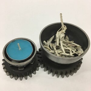 Puede incluir: Dos portavelas de metal en forma de engranaje. El portavelas más pequeño tiene una vela de té azul verdoso en su interior. El portavelas más grande tiene un cuenco con varillas de sahumerio de salvia blanca en su interior.