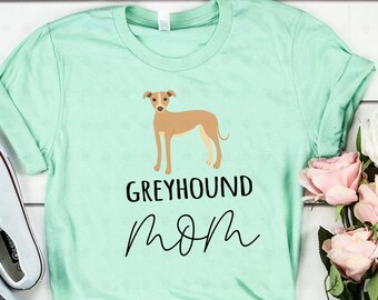 May include: A light green t-shirt featuring a brown and white illustration of a Greyhound dog. The text "Greyhound Mom" is printed in black below the dog.