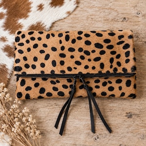 May include: A rectangular fold-over clutch bag featuring a tan and black leopard print pattern on a textured hair-on hide surface. The bag includes a black zipper closure with two long black leather zipper pulls hanging from the center.