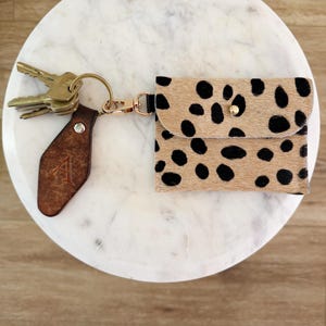 May include: A small rectangular pouch with a tan and black spotted animal print pattern, featuring a gold-tone snap closure. It is attached to a gold-tone keyring holding several metal keys and a dark brown leather tag embossed with the letter A.