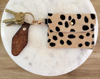 May include: A small rectangular pouch with a tan and black spotted animal print pattern, featuring a gold-tone snap closure. It is attached to a gold-tone keyring holding several metal keys and a dark brown leather tag embossed with the letter A.