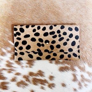 May include: A rectangular clutch wallet featuring a tan and black spotted animal print pattern on a textured hair-on-hide surface. The wallet has a fold-over flap closure secured with a single small, centered metallic stud fastener.