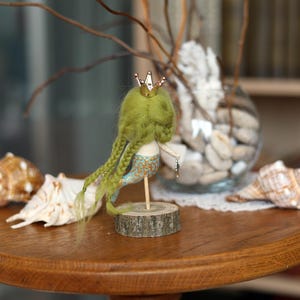 May include: A handmade mermaid doll with green hair and a gold crown, mounted on a wooden base. The mermaid has a blue and gold tail. Seashells and a glass vase with stones are in the background, all on a wooden table.