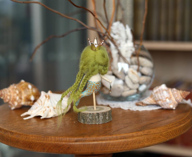 May include: A handmade mermaid doll with green hair and a gold crown, mounted on a wooden base. The mermaid has a blue and gold tail. Seashells and a glass vase with stones are in the background, all on a wooden table.