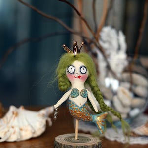 May include: A whimsical handmade mermaid doll with green yarn hair, a gold crown, and a blue and gold tail. The doll is mounted on a wooden base and has a painted face with large eyes and a red smile.