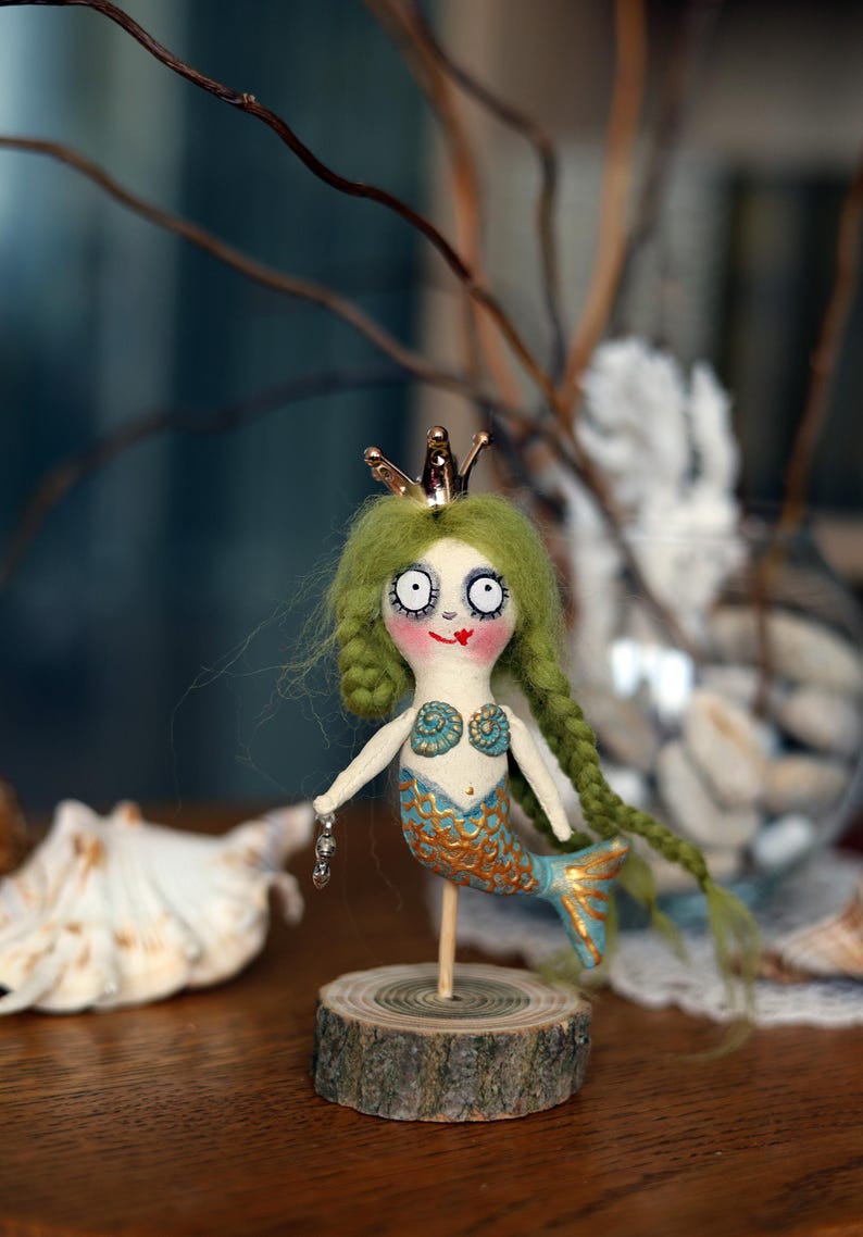 May include: A whimsical handmade mermaid doll with green yarn hair, a gold crown, and a blue and gold tail. The doll is mounted on a wooden base and has a painted face with large eyes and a red smile.