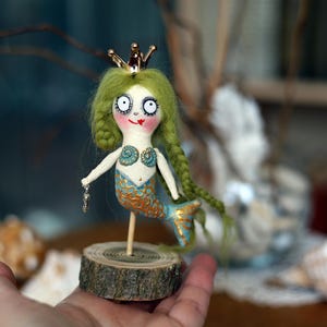 May include: A whimsical handmade mermaid doll with green yarn hair, a gold crown, and a blue and gold tail. The doll is mounted on a small wooden base and holds a small charm. The mermaid's face has large eyes and a painted smile.