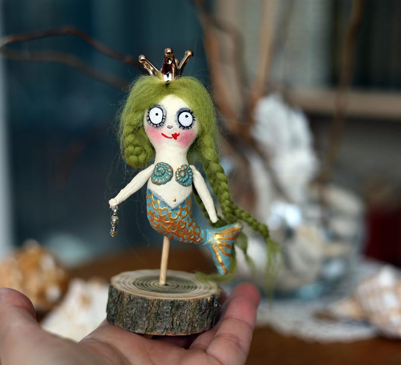 May include: A whimsical handmade mermaid doll with green yarn hair, a gold crown, and a blue and gold tail. The doll is mounted on a small wooden base and holds a small charm. The mermaid's face has large eyes and a painted smile.