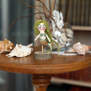 May include: A whimsical mermaid doll with green hair and a gold crown stands on a wooden base. The doll has a painted face and a blue and gold tail. Seashells and a glass vase with decorative elements are on a round wooden table.