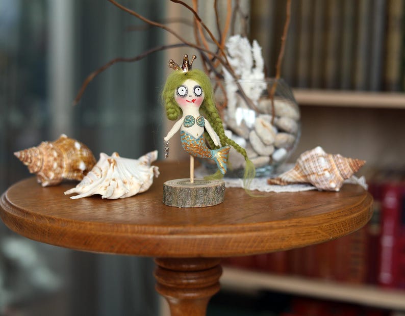 May include: A whimsical mermaid doll with green hair and a gold crown stands on a wooden base. The doll has a painted face and a blue and gold tail. Seashells and a glass vase with decorative elements are on a round wooden table.
